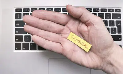 Uma mão na frente de um teclado de laptop, segurando um pedaço de papel com a senha escrita nele.