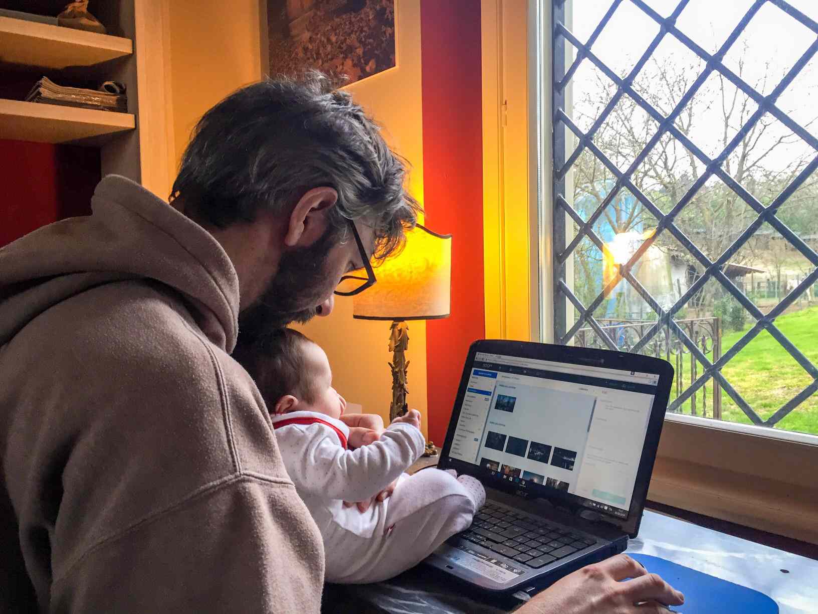 Um homem trabalhando em seu laptop em casa, segurando seu bebê.
