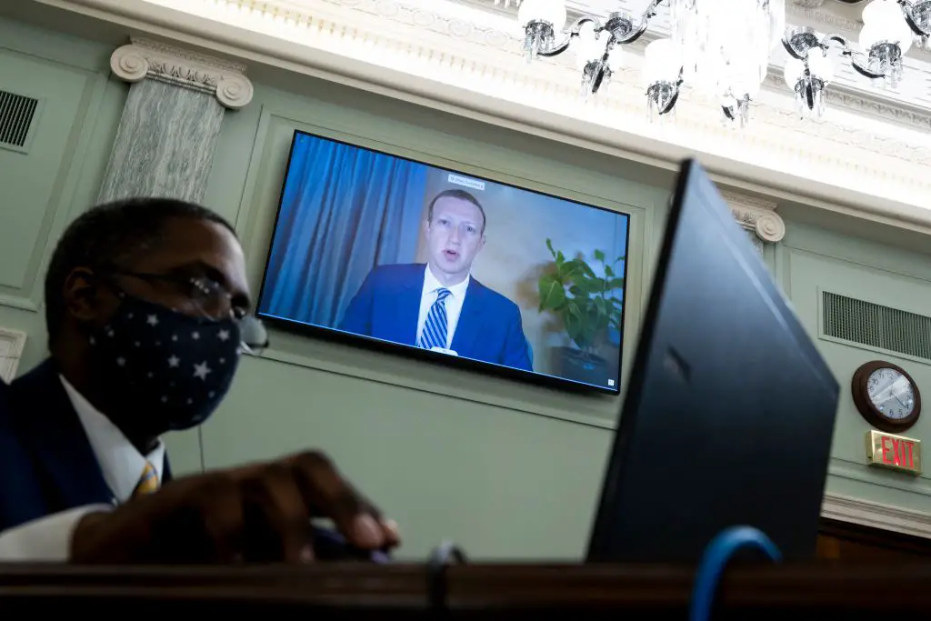 Mark Zuckerberg aparece em uma TV atrás de uma pessoa em uma mesa usando uma máscara de pano