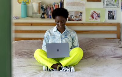 Uma mulher sentada de pernas cruzadas em sua cama enquanto usava um laptop Microsoft Surface.