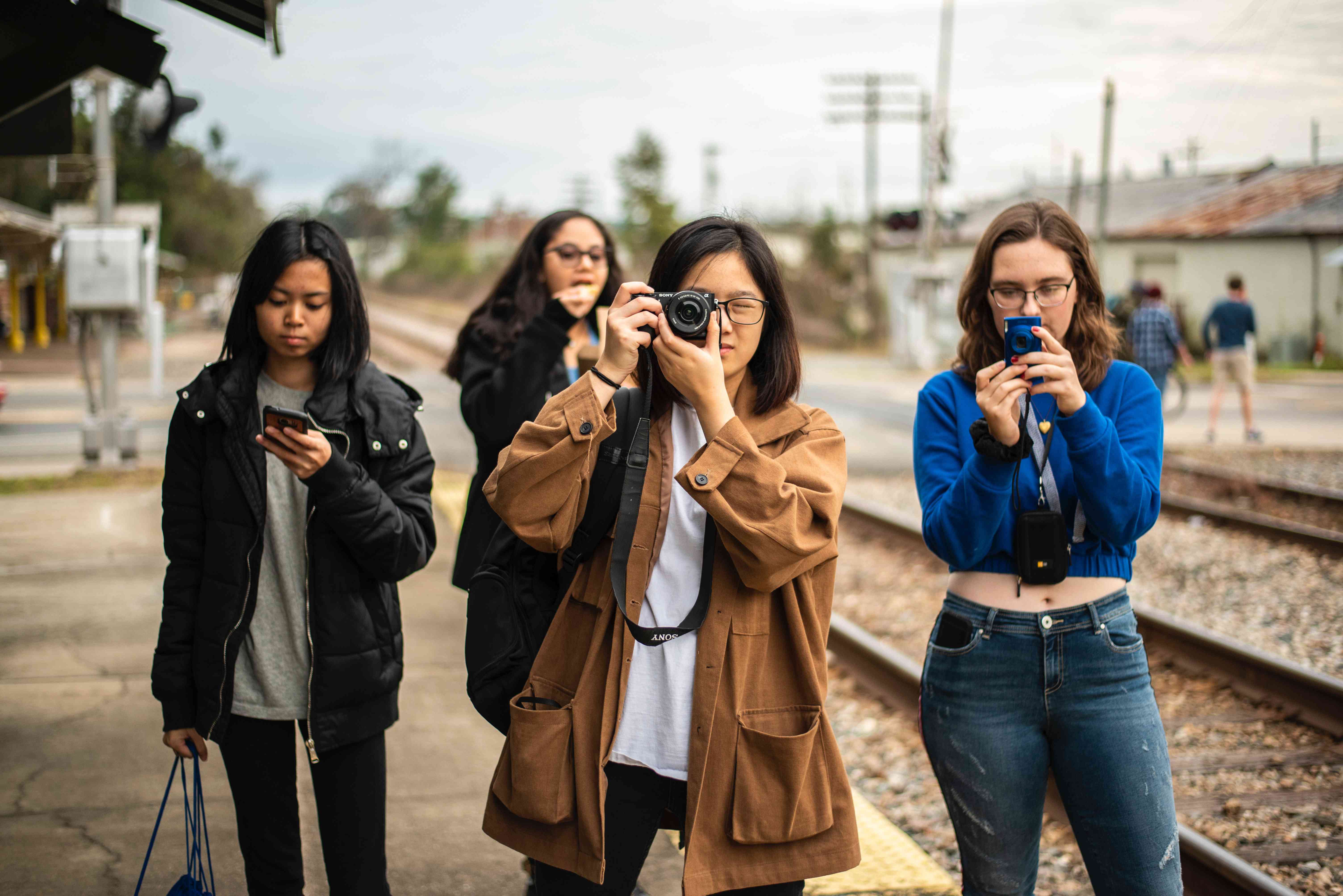 Quatro pessoas paradas perto dos trilhos da ferrovia, uma usando uma câmera sem espelho e outra uma câmera aponte e dispare. 