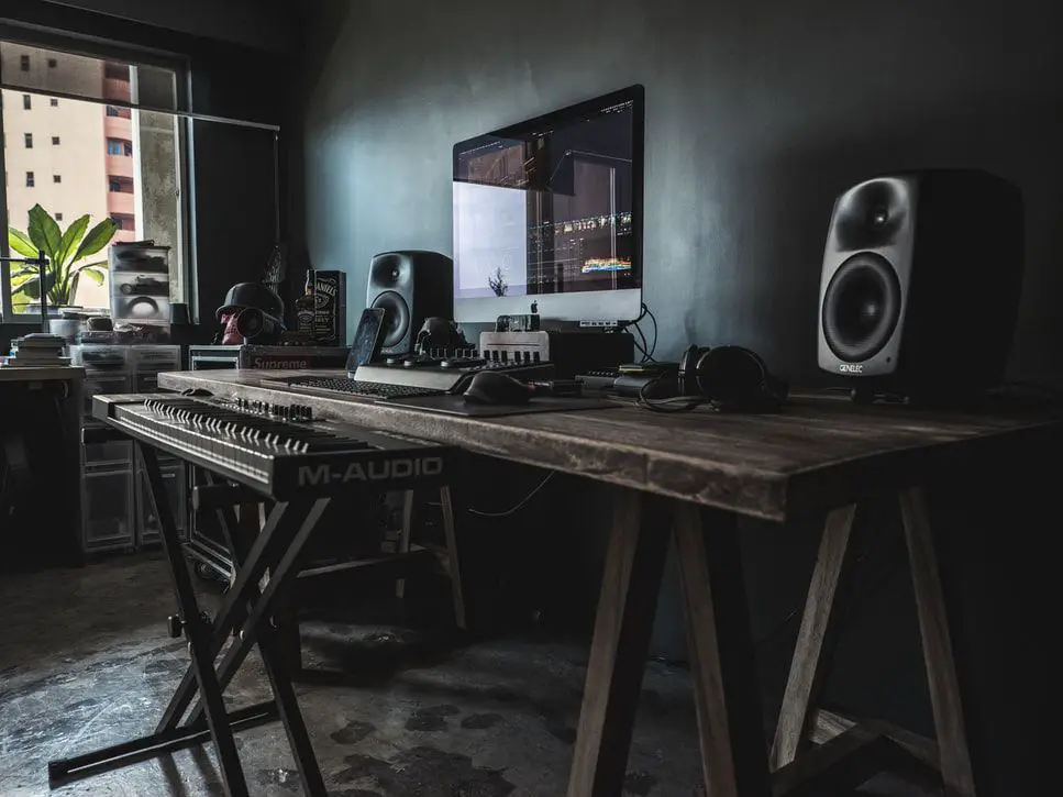 Mesa de madeira marrom com um computador Mac e um teclado musical em pé na frente dela.