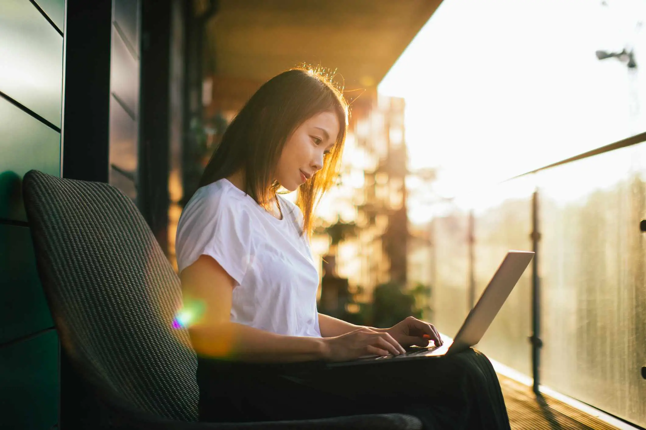 Mulher trabalhando com o computador no colo, sentada na varanda ao pôr do sol