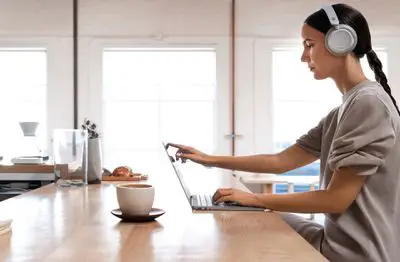 Uma mulher usando fones de ouvido Surface enquanto usava um Laptop Surface em sua cozinha.