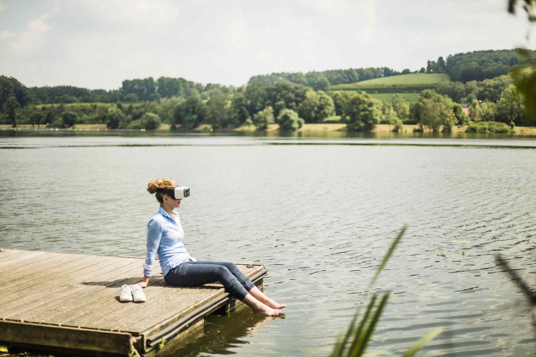 Alguém com óculos de realidade virtual sentado em um cais no lago com os pés na água. 