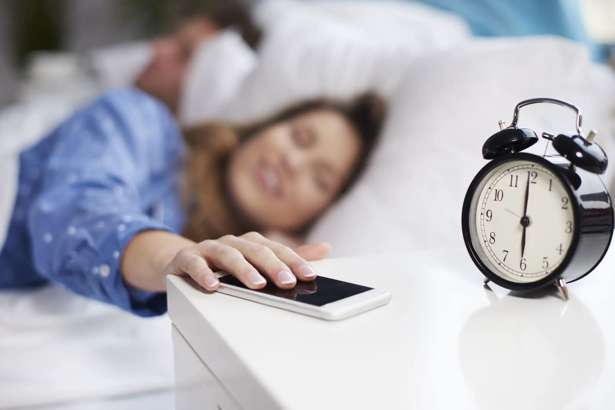 mulher na cama tocando seu telefone ao lado de um despertador