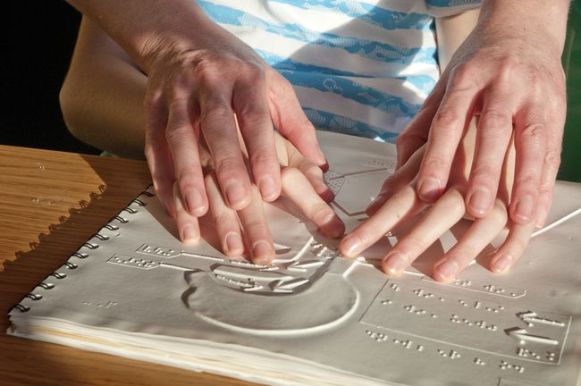 Closeup nas mãos de adultos orientando as mãos de uma criança sobre impressão de brail em um livro.