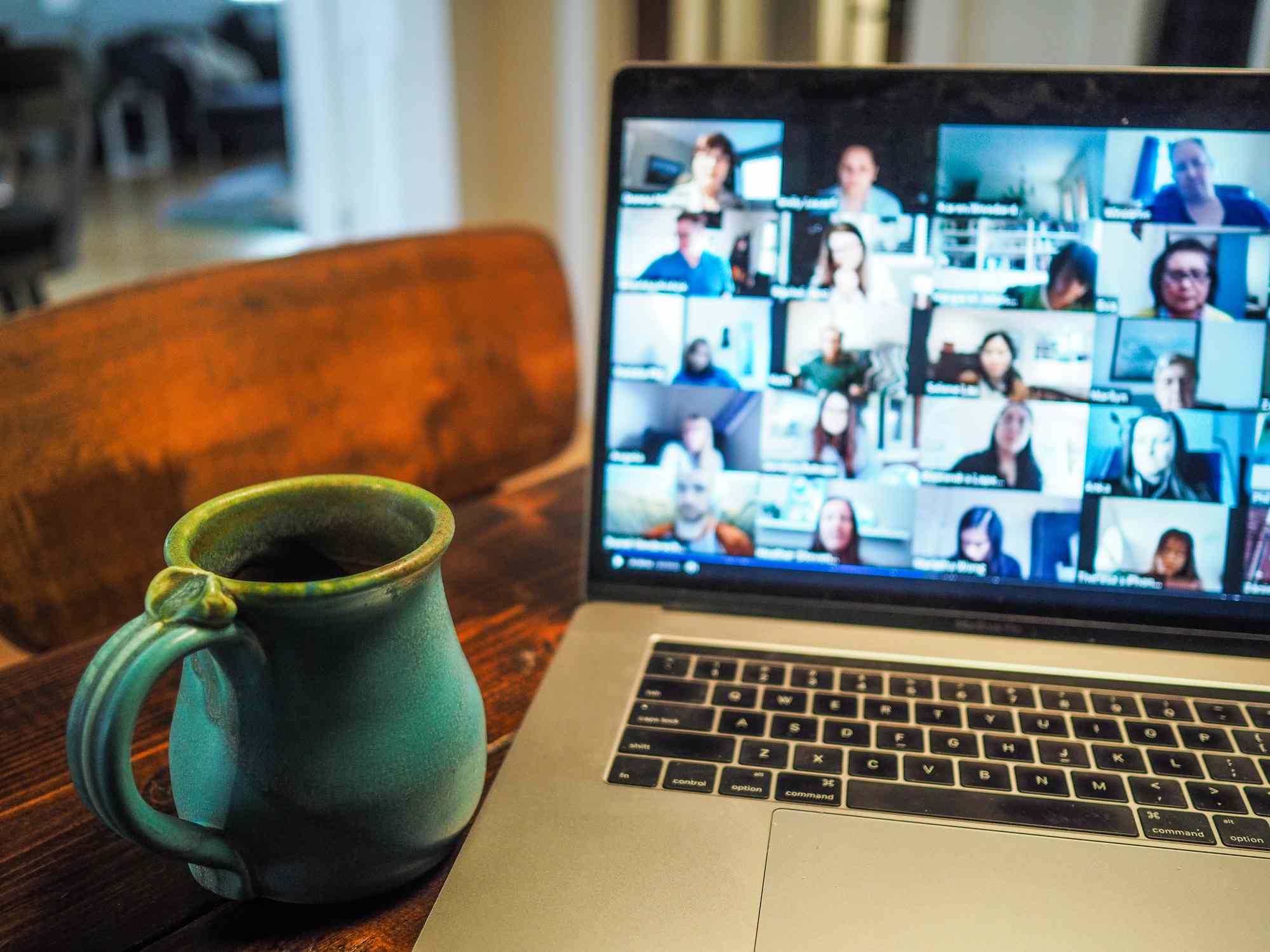 Um laptop aberto em uma mesa de cozinha exibindo uma videochamada com várias pessoas com uma xícara de café por perto. 