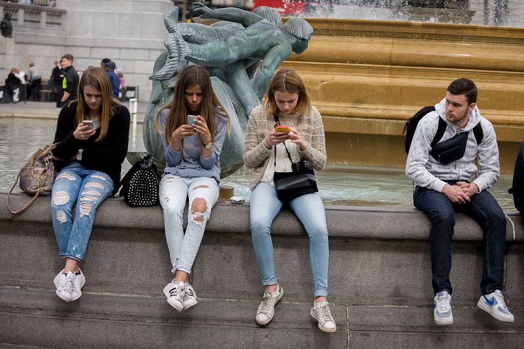 Três adolescentes estão perdidas em seus smartphones enquanto um jovem olha para elas 
